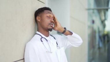 Hastanenin dışında dikilen stresli Afrikalı Amerikalı doktor kliniğin yakınında düşünceli bir doktor. Salgın sırasında steteskoplu sıhhiyeci doktor önlüğü giymenin stresi