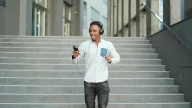 Genç, hareketli, Afro-Amerikan bir adam, kulaklıkla yürüyor, müzik dinliyor, açık havada şehir modern sokak arka planında. Komik adam dans ederken akıllı telefon kulaklığı kullanıyor.