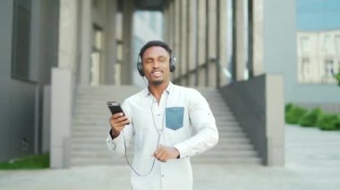 Genç, hareketli, Afro-Amerikan bir adam, kulaklıkla yürüyor, müzik dinliyor, açık havada şehir modern sokak arka planında. Komik adam dans ederken akıllı telefon kulaklığı kullanıyor.