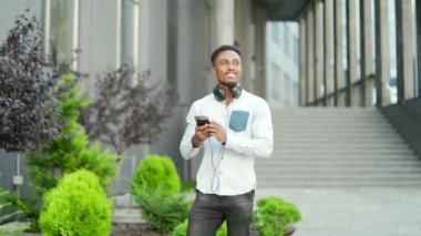 Neşeli Afro-Amerikan adam şehir caddesinde günlük kıyafetleriyle cep telefonu kullanarak yürüyor. Modern geçmişe sahip akıllı telefonlu bir erkek hippi sohbete gider. Gülümseyen mutlu erkek. Siyah adam.