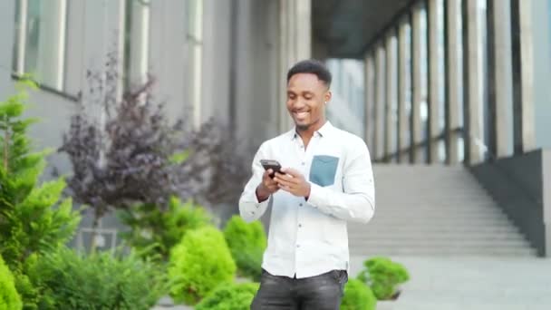 陽気なアフリカ系アメリカ人男性が携帯電話を使ってカジュアルな服装で都会の通りを歩いています 現代の背景に手にスマートフォンを持つ男性のヒップスターチャットに行きます 幸せな笑顔の男性 黒人男性 — ストック動画