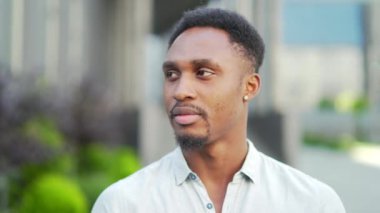 Dışarıda kameraya bakan ciddi bir Afro-Amerikalı portreyi yakından çek. Modern şehir caddesinde rahat gömlekli, kendine güvenen genç siyahi bir adamın yüzü arka planda bulanıklaştı. Yakın çekim