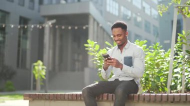 Mutlu Afro-Amerikan adam açık havada bankta oturuyor, elinde cep telefonuyla iyi haberler okuyor, dışarıda akıllı telefon sevinçlerini seyrederek zaferi kazanıyor. Erkek duyguları öğrenci sonuçları