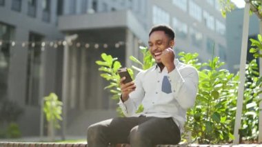 Mutlu Afro-Amerikan adam açık havada bankta oturuyor, elinde cep telefonuyla iyi haberler okuyor, dışarıda akıllı telefon sevinçlerini seyrederek zaferi kazanıyor. Erkek duyguları öğrenci sonuçları