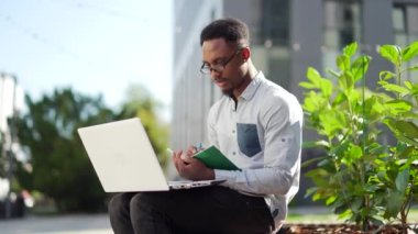 Genç Afro-Amerikan erkek öğrenci bir bankta dizüstü bilgisayarı ve defteriyle oturuyor. İnternette açık havada ders çalışıyor. Kitapları olan serbest çalışan bir adam sokak e-öğreniminde uzaktan öğreniyor.