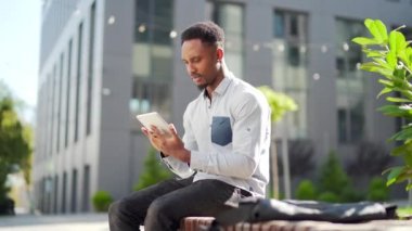 Afro-Amerikan adam tablet kullanıyor. Şehir sokaklarında kentin arka planında bankta oturuyor. Zenci adam elinde bir alet ya da akıllı telefon tutarak okuyor ya da alışveriş yapıyor. Kampüste bir öğrenci. Dışarıdaki serbest çalışan.