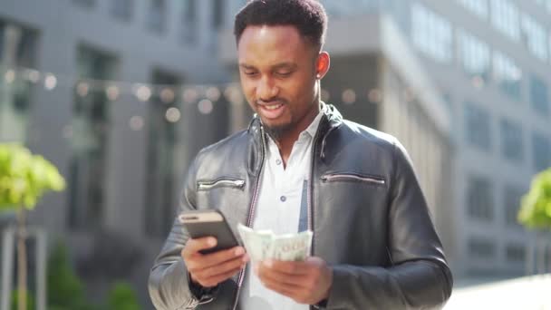 通りの近代的な建物の上に立つアフリカ系アメリカ人の男は手に携帯電話で現金お金を賭けることで収益を示しています 幸せな黒男屋外でスマートフォンとパックドル — ストック動画