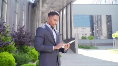 Resmi takım elbiseli genç Afro-Amerikan iş adamı dışarıda arka plandaki modern ofis binasında elinde tabletle çalışıyor. Şehir dışında akıllı telefon ya da cep telefonu kullanan bir adam.