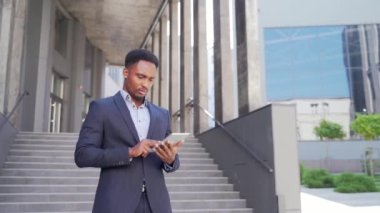 Resmi takım elbiseli genç Afro-Amerikan iş adamı dışarıda arka plandaki modern ofis binasında elinde tabletle çalışıyor. Şehir dışında akıllı telefon ya da cep telefonu kullanan bir adam.