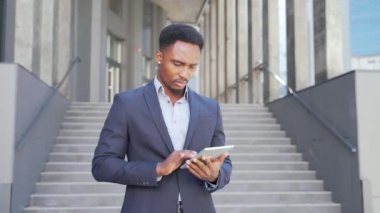 Resmi takım elbiseli genç Afro-Amerikan iş adamı dışarıda arka plandaki modern ofis binasında elinde tabletle çalışıyor. Şehir dışında akıllı telefon ya da cep telefonu kullanan bir adam.