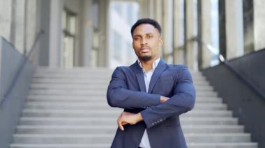 Genç özgüvenli Afro-Amerikan iş adamının modern ofis binasının arka planında duran ciddi iş adamına baktığı portre. Erkek, kollarını kavuşturmuş resmi bir takım elbise.