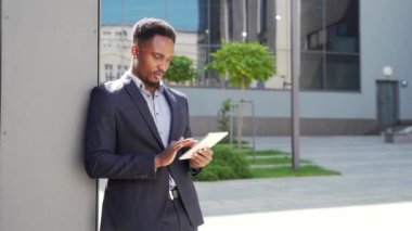 Resmi takım elbiseli genç Afro-Amerikan iş adamı dışarıda arka plandaki modern ofis binasında elinde tabletle çalışıyor. Şehir dışında akıllı telefon ya da cep telefonu kullanan bir adam.