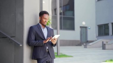 Resmi takım elbiseli genç Afro-Amerikan iş adamı dışarıda arka plandaki modern ofis binasında elinde tabletle çalışıyor. Şehir dışında akıllı telefon ya da cep telefonu kullanan bir adam.