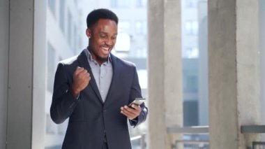 Smartphone 'a bakarak başarıyı kutlayan Afrikalı Amerikalı iş adamı çıktı. Başarılı bir iş adamı zaferi dışarıda cep telefonuyla kazanır. Mutlu İyi Haberler Başarı Anlaşması, Başlangıç Bahis Kazanma oranı