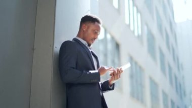 Resmi takım elbiseli genç Afro-Amerikan iş adamı dışarıda arka plandaki modern ofis binasında elinde tabletle çalışıyor. Şehir dışında akıllı telefon ya da cep telefonu kullanan bir adam.
