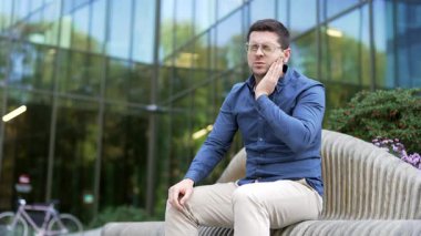 İş yeri binasının yanındaki bankta oturan diş ağrısından muzdarip iş adamı. Üzgün, yetişkin, yakışıklı bir adam elini yanağında tutuyor. Yüzünde masaj izi var. Dişçide tedavi olması gerekiyor.