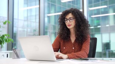 Kendine güvenen iş kadını, modern cam iş ofisindeki çalışma masasında otururken dizüstü bilgisayarda çalışıyor. Bilgisayarda yazan günlük gömlekli kadın çalışan, görevlerle meşgul olan bir projeyle meşgul.