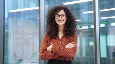 Modern cam iş ofisindeki iş yerinde kollarını kavuşturmuş sıradan gömlekli gülümseyen iş kadınının portresi. Kameraya bakarak poz veren mutlu kadın yönetici ya da çalışan.