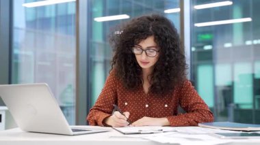 İş kadını, modern iş ofisindeki bir masada oturan belgeleri imzalıyor. Kendine güveni tam bir kadın yönetici. Dosyayı evraklarla karıştırıyor, kontrol ediyor ve sözleşmeyi kalemle imzalıyor.