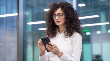 İş kadını, modern iş ofisindeki iş yerinde dururken telefonu kullanıyor. Kadın yönetici mobil uygulama üzerinde çalışıyor, daktilo mesajı okuyor, internette sohbet ediyor ya da akıllı telefondan internette geziniyor. Kapat.