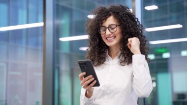 Modern bir cam şirketinde dururken cep telefonundan iyi haberler okuyan memnun iş kadını. Kadın çalışanın mutlu olması akıllı telefondan olumlu mesaj iletiyor.