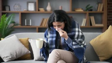 Evdeki oturma odasında kanepede oturan soğuk algınlığı ve gripten muzdarip hasta genç kadın. Sağlıksız yetişkin bir dişi battaniyeye sarılmış öksürük, burnunu mendille siler ve sıcak çay içer.