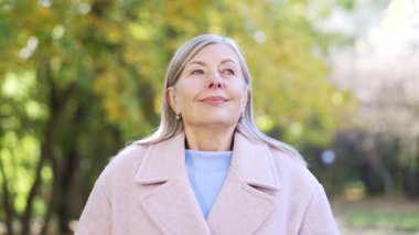 Mutlu bir portre, yaşlı, gri saçlı, derin nefes alan, gülümseyen ve sonbahar parkında palto giymiş ağaçların arasında kameraya bakan bir kadın. Kafasından vurulmuş yaşlı bir kadın doğada rahatlar.