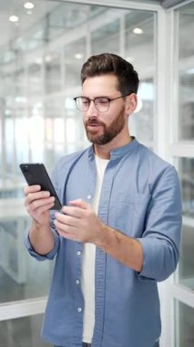 Memnun iş adamı iş yerindeki cep telefonundan harika haberler aldı. Gülümseyen yakışıklı girişimci akıllı telefondan olumlu bir mesaj okur. Dikey video