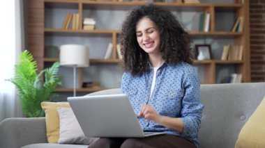 Evdeki oturma odasındaki kanepede oturup dizüstü bilgisayardan harika haberler okuyan mutlu genç bir kadın. Kadın serbest çalışanın bilgisayardan olumlu bir mesaj almasına sevinen, başarıya sevinen