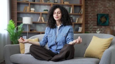 Evde oturma odasında kanepede otururken nilüfer pozisyonunda, gözleri kapalı meditasyon yapan sakin genç bir kadın. Mutlu güzel bayan yoga yapar, derin derin nefes alır, huzuru hisseder, kanepede dinlenir.