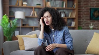 Evdeki oturma odasında kanepede otururken baş ağrısı çeken sağlıksız genç bir kadın. Yetişkin olmayan dişi, şakaklarına elleriyle masaj yapıyor. Mutsuz kız acı çekiyor, yüksek tansiyonu ya da migreni var.