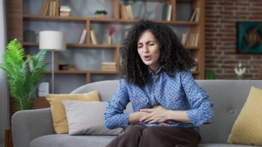 Evdeki oturma odasında kanepede oturan, karın ağrısından muzdarip hasta genç bir kadın. Sağlıksız kadın ellerini karnında tutuyor. Zehirlenme, kabızlık, şişkinlik ya da adet görme.