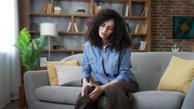 Sağlığı yerinde olmayan genç bir kadın evdeki oturma odasında kanepede otururken şiddetli diz eklem ağrısı çekiyor. Üzgün yetişkin kadınlar bacak kaslarına masaj yapar, yürüyemez, hastalık, kramp veya romatizma geçiremez.