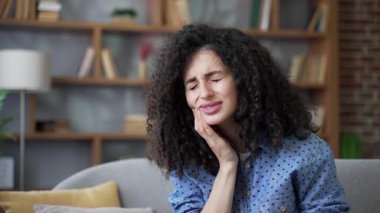 Evdeki oturma odasındaki kanepede oturan hasta genç kadın diş ağrısından muzdarip. Üzgün kadın, elini yanağında tutuyor ve masaj yapıyor. Keskin bir ağrı hissediyor, dişçiye görünmesi gerekiyor. Kapat.