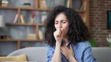 Evdeki oturma odasında kanepede oturan hasta genç kadın öksürüyor. Sağlıksız yetişkin bir dişi kendini kötü hisseder, göğüs göğse tutuşur. Hasta kadın nezle, virüs, grip, bronşit ya da zatürree olmuş. Kapat.