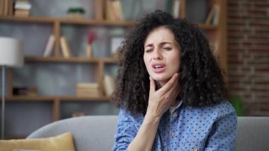 Evdeki kanepede oturan hasta, boğazı ağrıyan genç bir kadın. Sağlıksız yetişkin bir kadın soğuk algınlığı, virüs ya da grip olabilir. Acı dolu bir ifadeyle boğaza dokunan bir kadın, masaj yapan hassas bir nokta. Kapat.