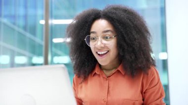 Mutlu Afro-Amerikan iş kadını laptopta harika haberler okuyor modern iş ofisindeki masasında oturuyor. Neşeli siyah kadın başarıyı kutluyor, gülümsüyor. Kapat.