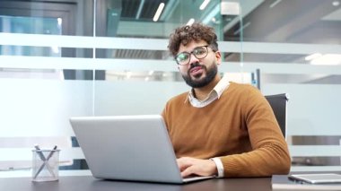 Modern cam işyerindeki bir masada oturan gülümseyen iş adamının portresi. Dizüstü bilgisayarda çalışan ve kameraya bakan kendinden emin başarılı bir erkek profesyonel.