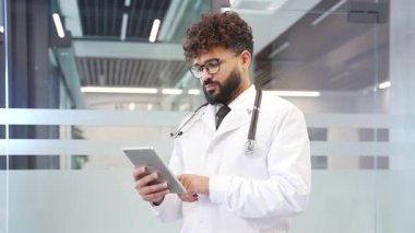 Modern hastane ofisinde dijital tablet kullanan beyaz önlüklü doktor. Sakallı tıp doktoru hastayla internette sohbet ediyor, internette bilgi tarıyor ya da başvuruda çalışıyor.