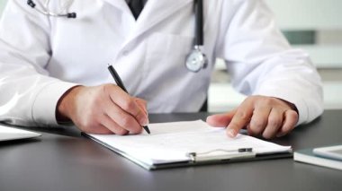 Hastane ofisindeki tıbbi belgeleri imzalarken yakın çekim. Laboratuvar önlüklü bir doktor pano, reçete, hasta kayıtları ya da tedavi sonuçlarıyla ilgili bir raporu inceliyor.