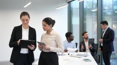 İki kadın çalışan ellerinde bir belge ve tablet tutarak iş ofisindeki çeşitli takım çalışanları üzerinde duruyorlar. İş arkadaşları iş konularını olumlu atmosferde tartışıyor