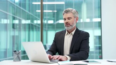 İş yerindeki çalışma masasında resmi takım elbiseli ciddi bir iş adamının portresi. Başarılı, sakallı, beyaz saçlı menajer laptopta çalışıyor ve kameraya bakıyor.