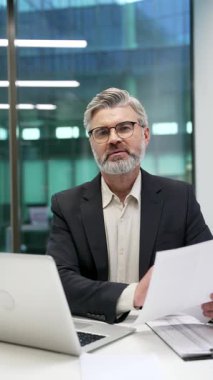 İş yerindeki masasında oturan resmi takım elbiseli olgun iş adamının portresi. Kendine güveni tam bir finansör. Evrak işleriyle meşgul. Bilgisayarda çalışıyor ve kameraya bakıyor. Dikey video