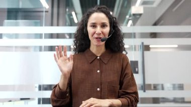 Webcam görüntüsü. Ofiste oturan kameraya bakan, kulaklıklı, gülümseyen bir iş kadını. Kadın yönetici çevrimiçi bir konferansta iletişim kurar ya da iş ortağıyla uzaktan sohbet eder