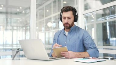 Kulaklıklı bir iş adamı iş yerinde iş yerinde otururken dizüstü bilgisayar kullanarak video konferansı izliyor. Güvenilir yönetici çevrimiçi toplantıda uzaktan iletişim kurar