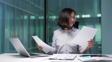 Neşeli Asyalı iş kadını, elinde tuttuğu belgelerle yürüttüğü mali raporun sonuçlarından memnun. Kadın müdür olumlu göstergelerden memnun.