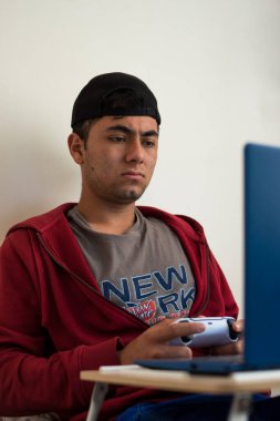 Kırmızı ceketli genç adam evdeki mavi dizüstü bilgisayarında beyaz bir kumandayla oyun oynuyor. 