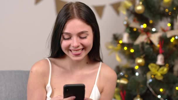 Woman during Christmas holidays stay home. Chat with someone trough smartphone. Look at camera and smile. Chatting with boyfriend — Stock Video