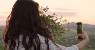 Genç bir kız telefonuyla dağlarda kır çiçekleriyle fotoğraf çekiyor.