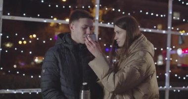 Genç adam, şehirde soğuk bir yürüyüş yaparken sevgilisinin elinden sıcak çay içer.